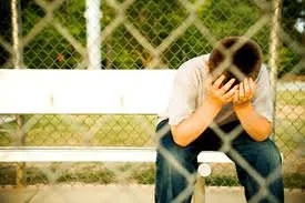 Sad boy sitting on a park bench behind a chainlink fence